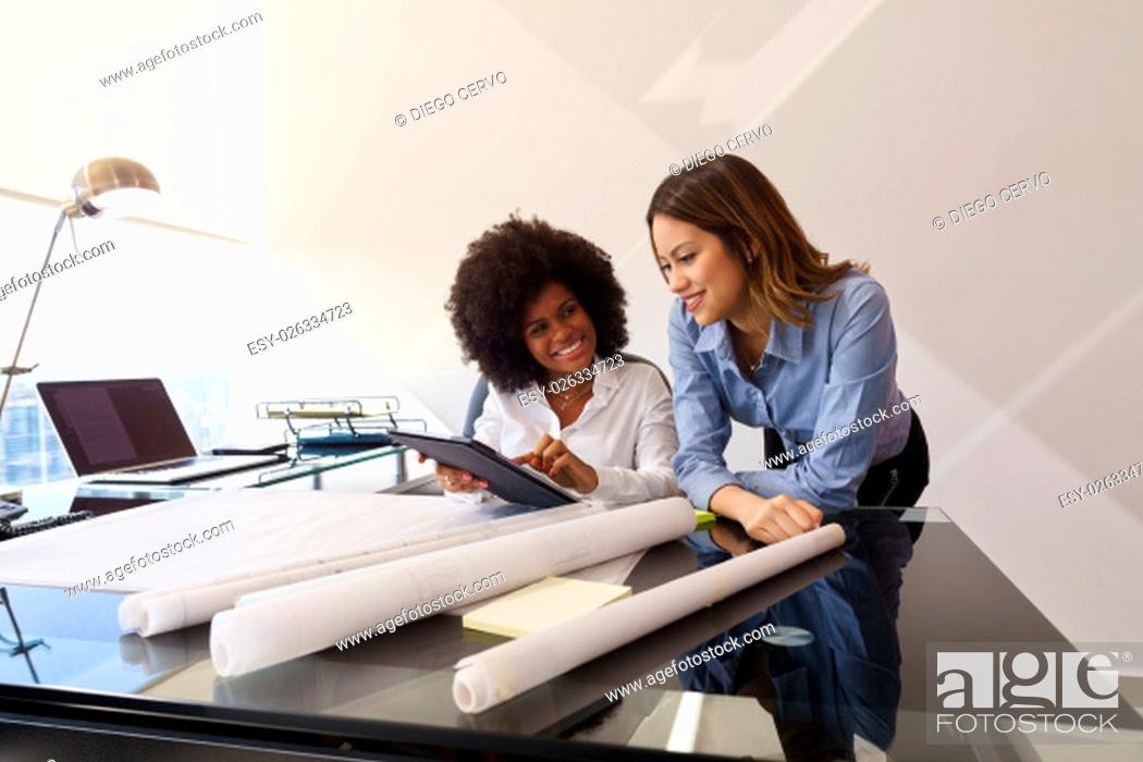 Stock Photo: Two architects in modern office building, sitting on desk with blueprints and housing projects. The women hold a tablet and surf the web.