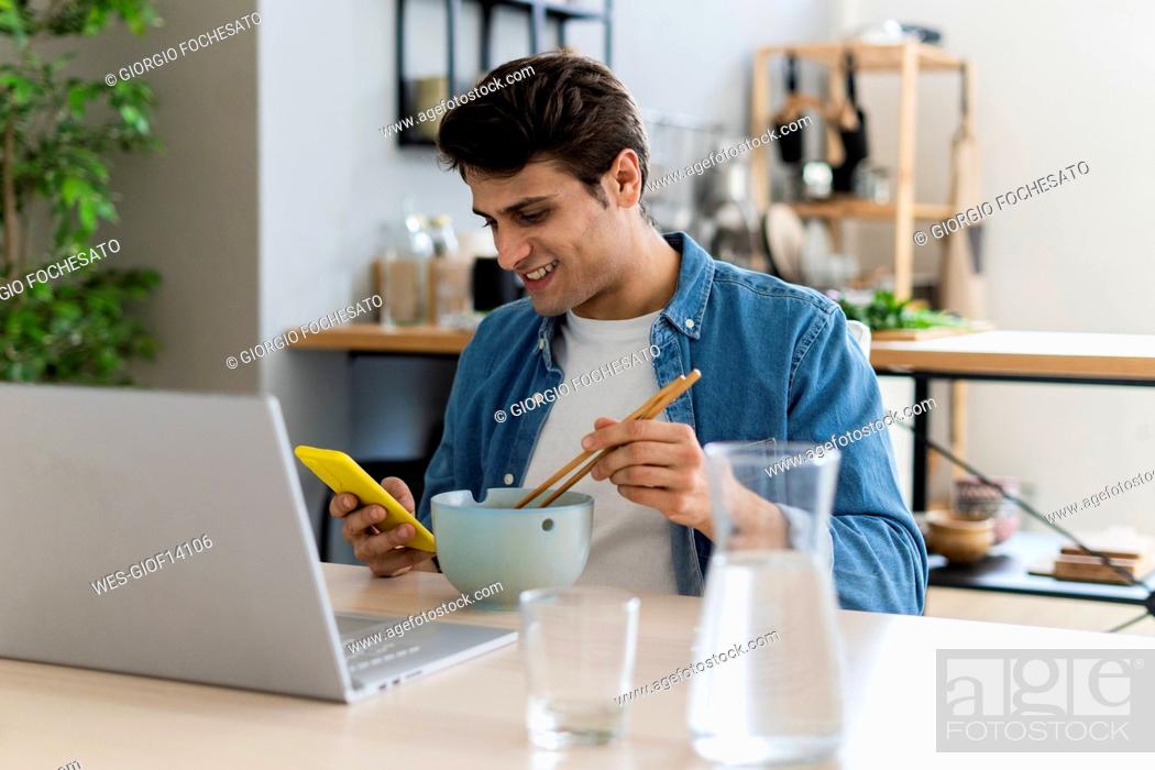 Stock Photo: Smiling man using mobile phone while having food at home.