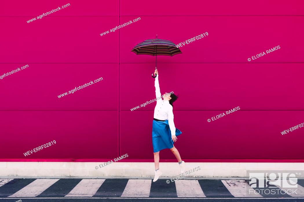 Stock Photo: Young woman jumping with umbrella in front of a pink wall.