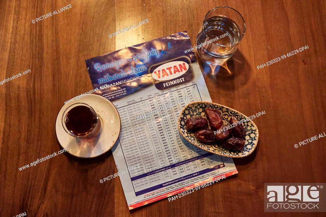 Stock Photo: 23 March 2023, Hamburg: A glass of water, a glass of tea, a bowl of dates and the Ramadan calendar with the times are on the table at the Pamukkale Köz.