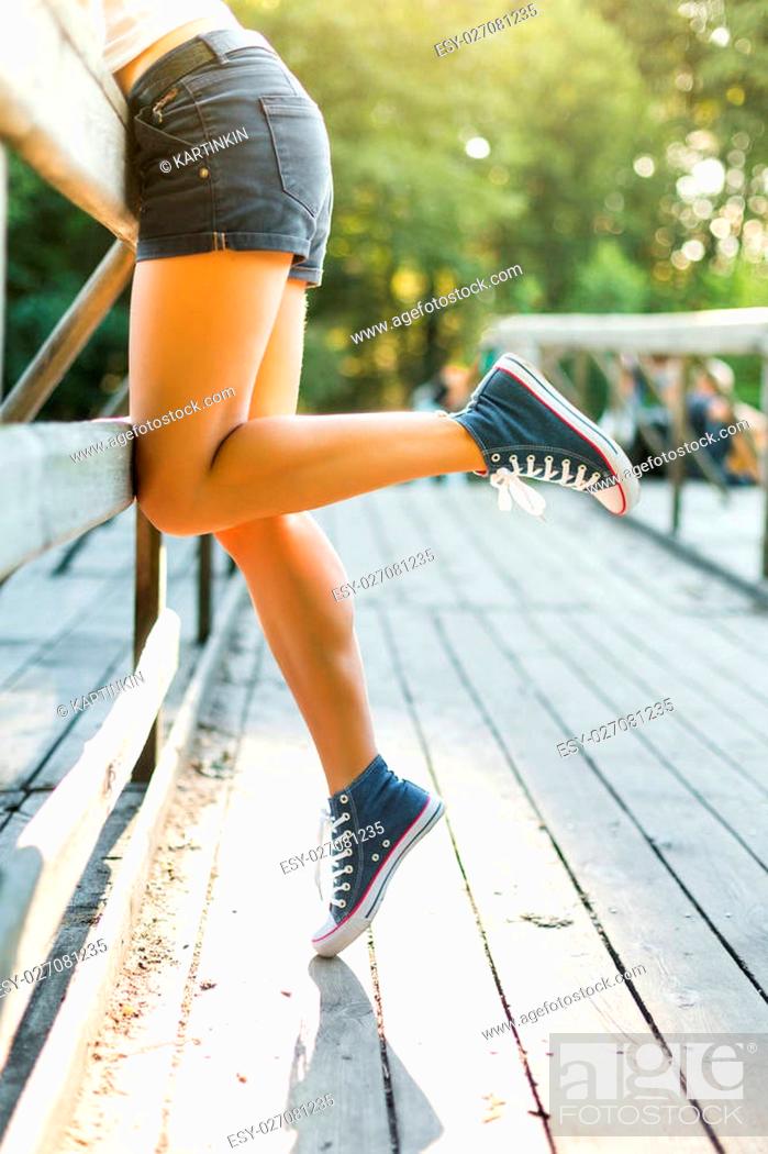 Stock Photo: Young woman with beautiful sporty legs leaning on a wooden bridge railing in jeans sneakers.