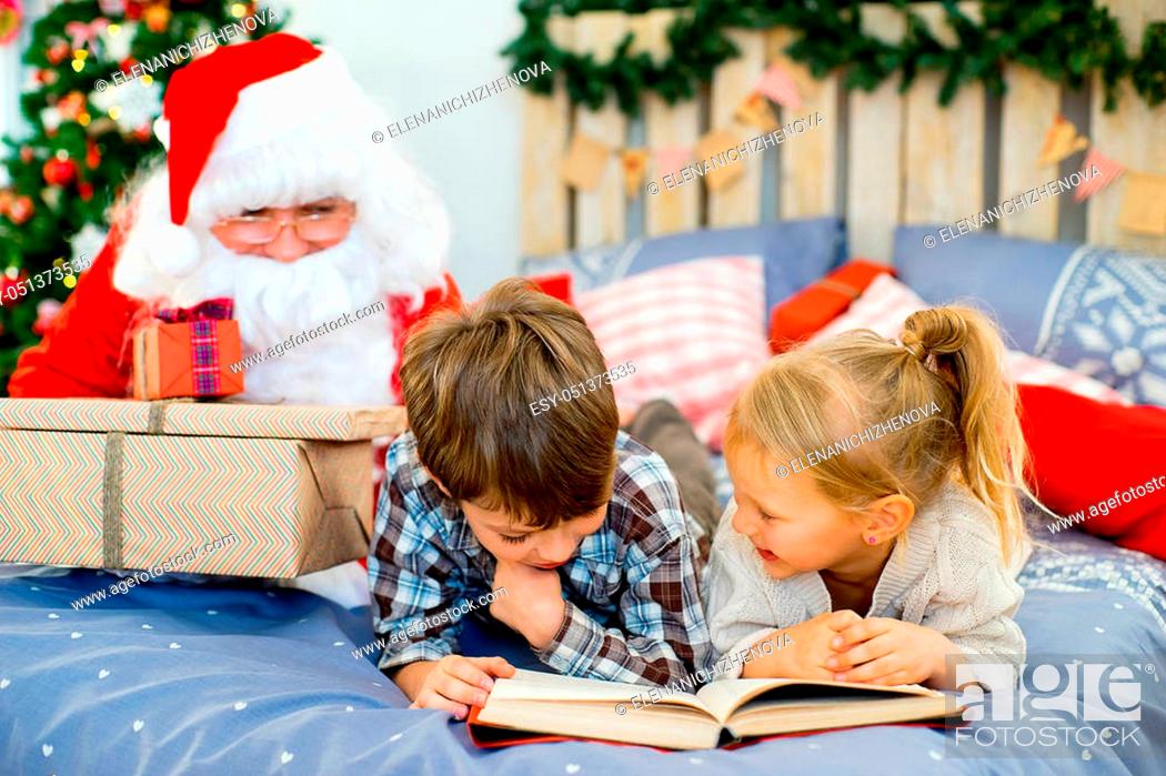 Stock Photo: Santa Claus quietly came to the children who are reading a book while lying on decorated bed with Christmas tree in the background.