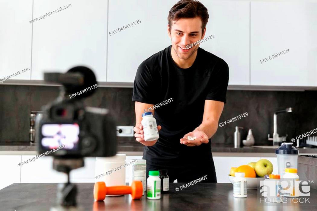 Stock Photo: Cheerful young man filming his video blog episode about healthy food additives while standing at the kitchen table and showing pills.