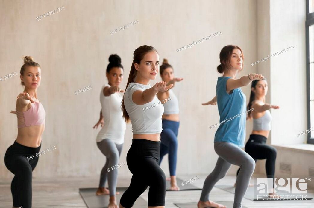 Stock Photo: Group of young sporty people practicing yoga lesson, doing Warrior Two pose, Virabhadrasana 2 exercise, working out, indoor full length.