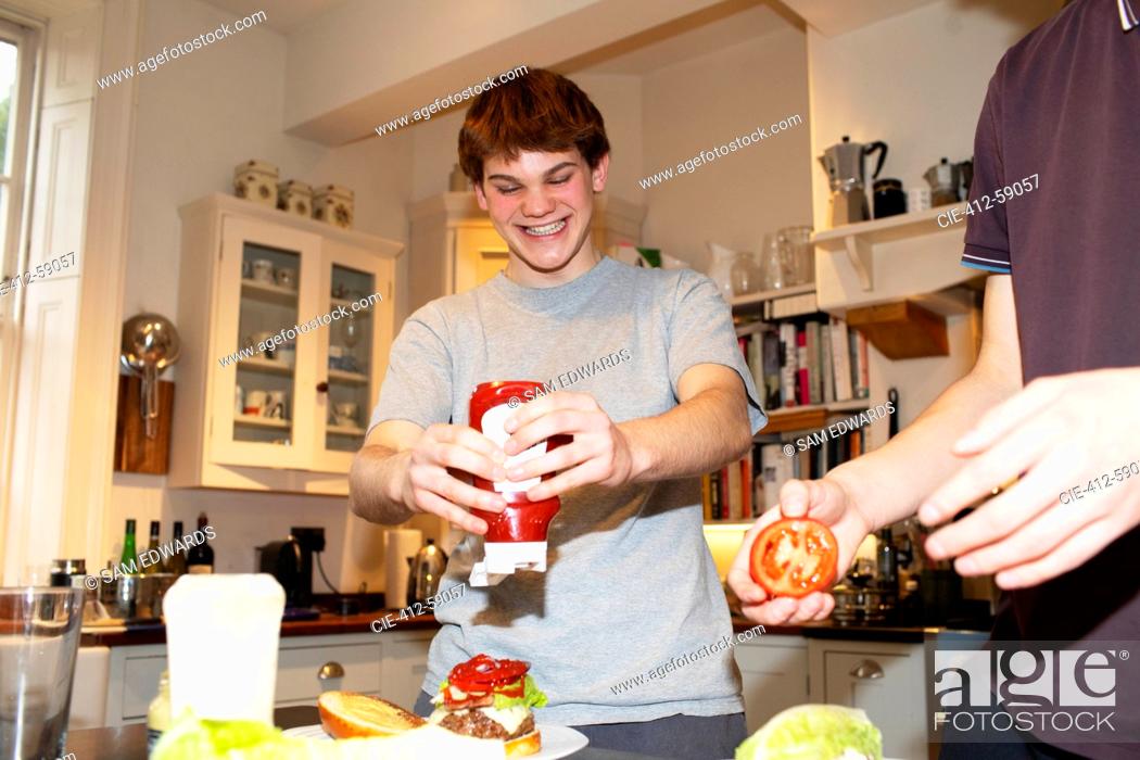 Imagen: Happy teenage boy squeezing ketchup onto hamburger in kitchen.