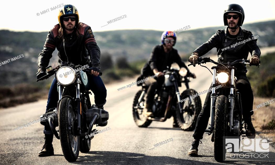 Stock Photo: Three men wearing open face crash helmets and goggles sitting on cafe racer motorcycles on a rural road.