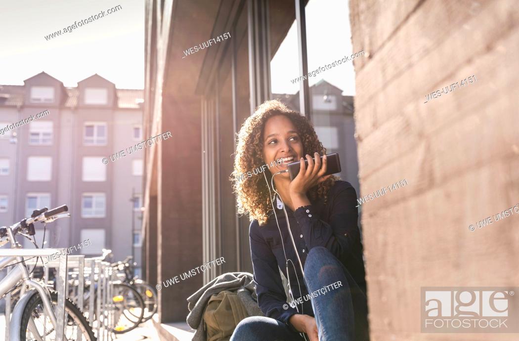 Stock Photo: Young woman sitting in front of window in the city, using smartphone.