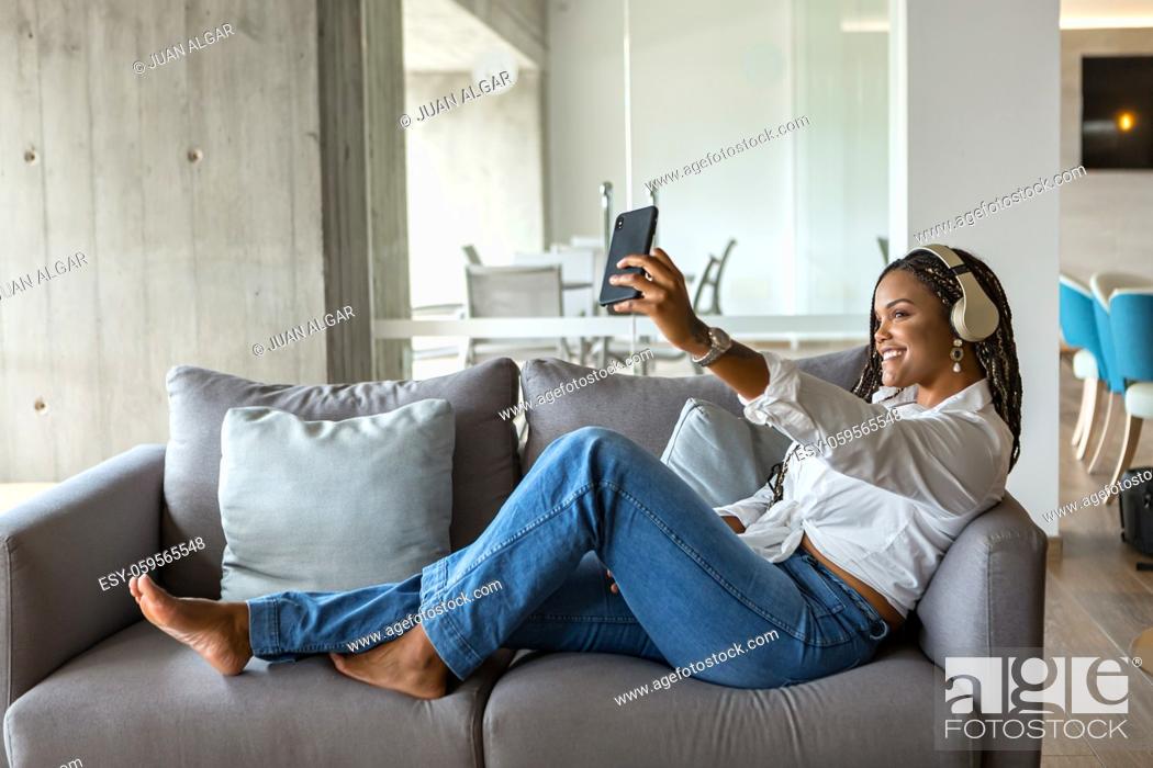 Imagen: Portrait of a happy young woman listening to music with headphones and taking a selfie while leaning on a sofa at home. Concept of people in home.