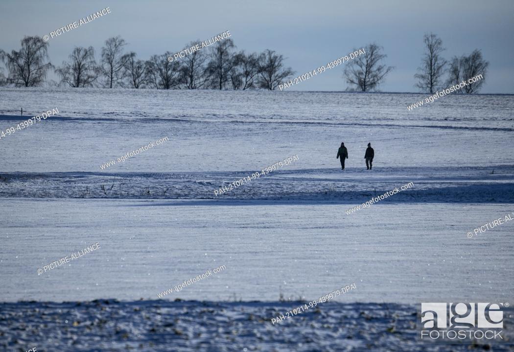 Stock Photo: 14 February 2021, Hessen, Dannenrod: Two walkers are walking along a country lane through the snow-covered landscape. The temperatures drop far below freezing.