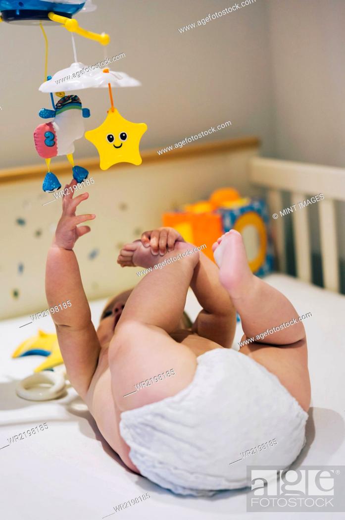 Stock Photo: A baby girl in a diaper lying in a cot reaching up to a colourful mobile hanging above her.