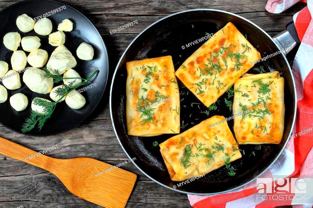 Stock Photo: delicious mozzarella spinach stuffed crepes wraps on skillet sprinkled with finely chopped dill, on dark wooden board with mini mozzarella balls on black plate.