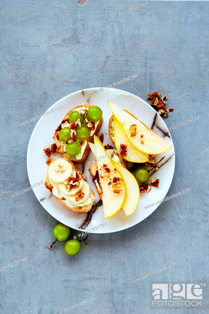 Stock Photo: Tasty crostini with pear, cream-cheese, grapes, banana and nuts. Top view of breakfast toasts or snack sandwiches on ceramic plate over grey stone background.