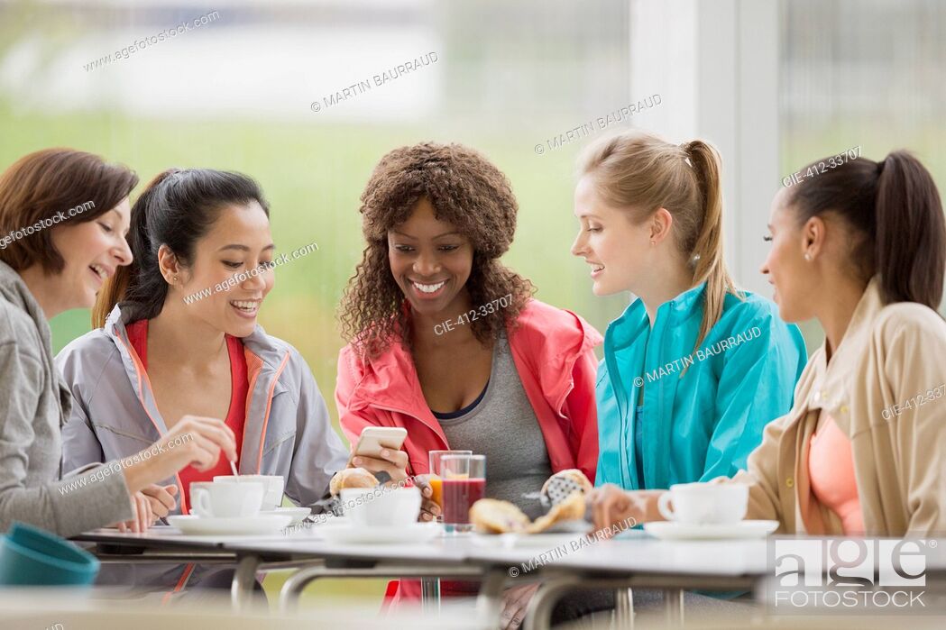 Imagen: Smiling women friends drinking coffee and using cell phone post workout.