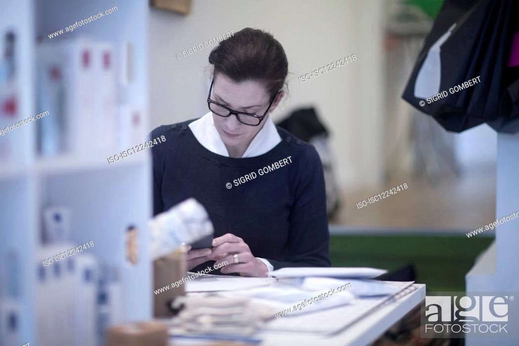 Stock Photo: Female tailor wearing glasses, sitting at table, checking her mobile phone.