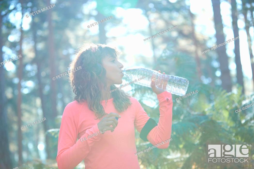 Stock Photo: Young female runner drinking bottled water in sunlit forest.