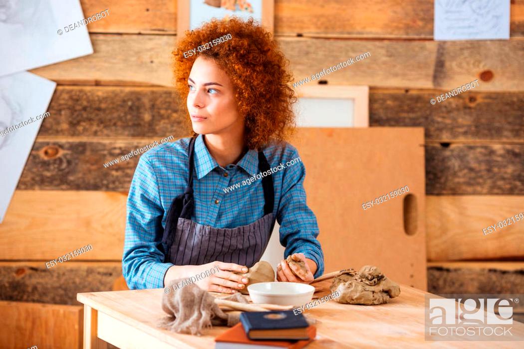 Stock Photo: Thoughtful curly young woman sitting in pottery workshop and making sculpture with clay.