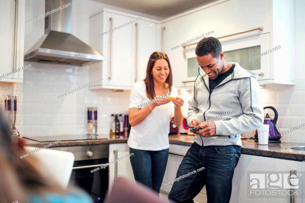 Stock Photo: Mid adult couple are relaxing in the kitchen of their home together. The woman is eating a slice of orange and the man is using his smart phone.