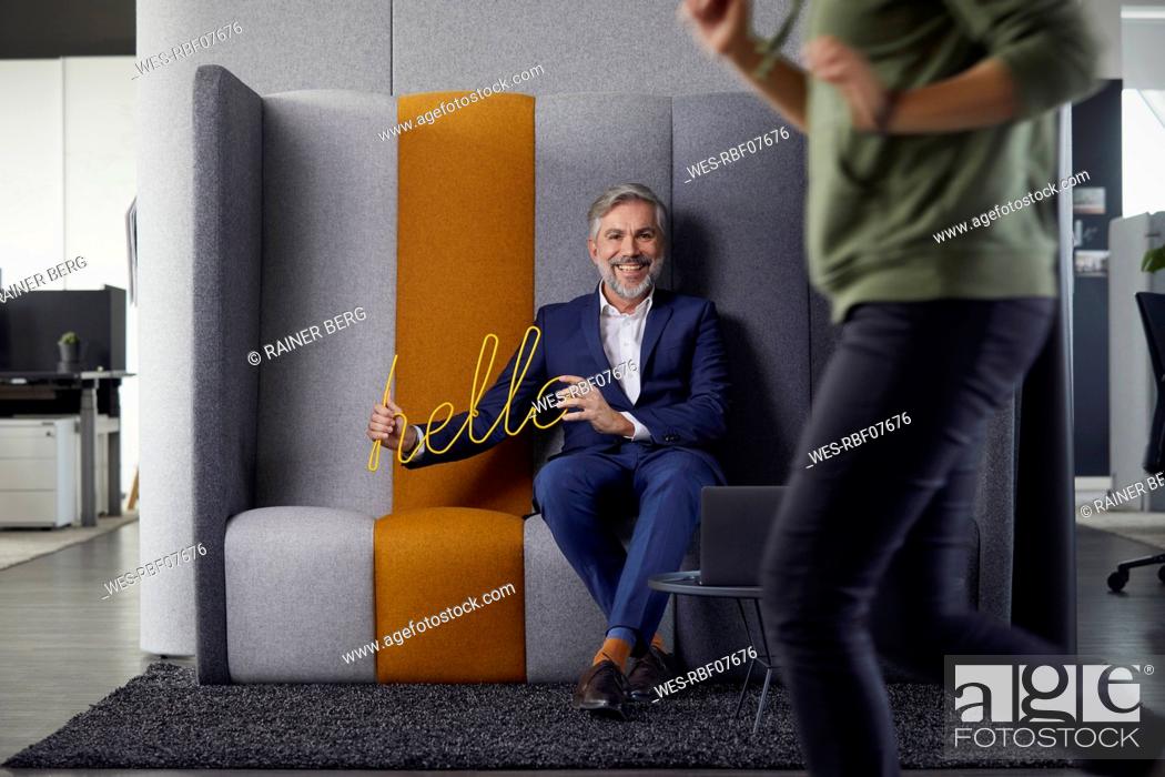 Stock Photo: Portrait of smiling mature businessman sitting on couch in office holding hello sign.