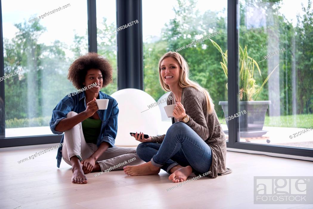 Stock Photo: Two young laughing multiethnic women sit on the floor near the window and enjoy while drinking coffee using smartphone in luxury home.