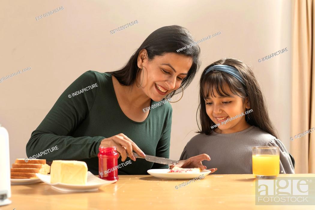 Stock Photo: AN ADULT WOMAN SPREADING JAM ON BREAD FOR HER DAUGHTER SITTING BESIDE.