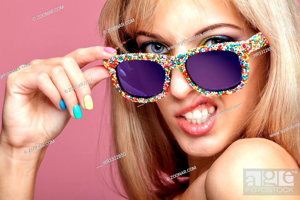 Imagen: Young blonde woman with fun candy glasses on pink background. Portrait of female in lilac dress with blue and yellow eyes makeup making grimace.
