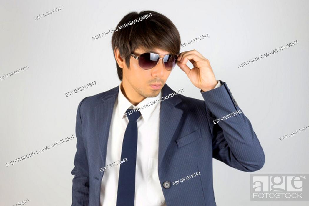 Photo de stock: Young Asian Portrait Businessman in Navy Blue Suit Touch Sunglasses and Look Beside on Grey Background.
