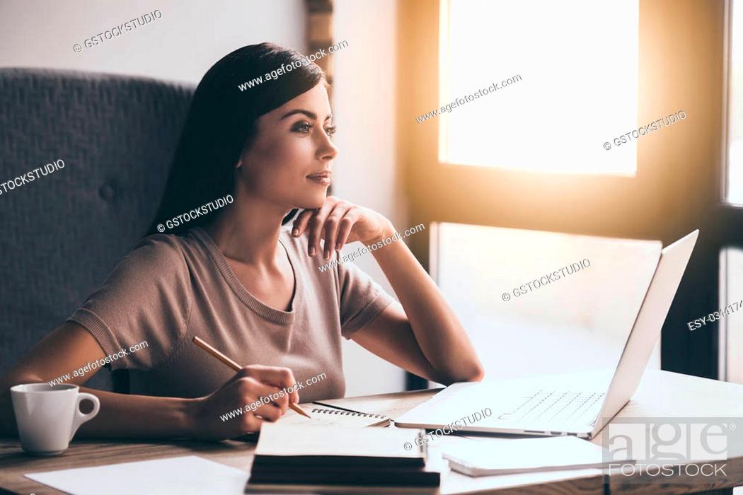 Stock Photo: Looking for inspiration. Young beautiful pensive woman making some notes and looking through window while sitting in chair at her working place.