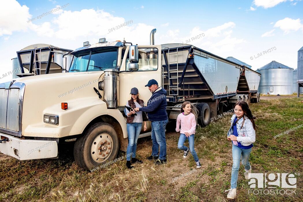 Stock Photo: Mature couple working on their farm, standing next to a diesel transport truck and consulting their tablet computer while their two daughters have fun running.
