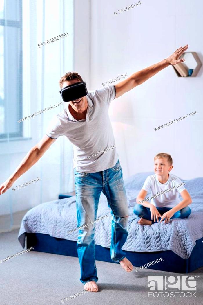 Imagen: New experience. Joyful young man standing wearing a VR headset, standing on one leg and spreading hands as if imitating flight while his son looking at him.