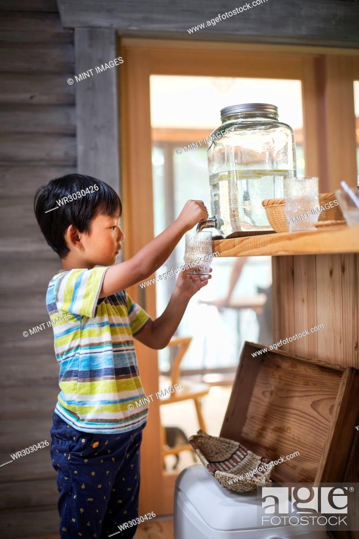 Stock Photo: Boy with black hair wearing stripy T-shirt filling drinking glass with water from water dispenser.