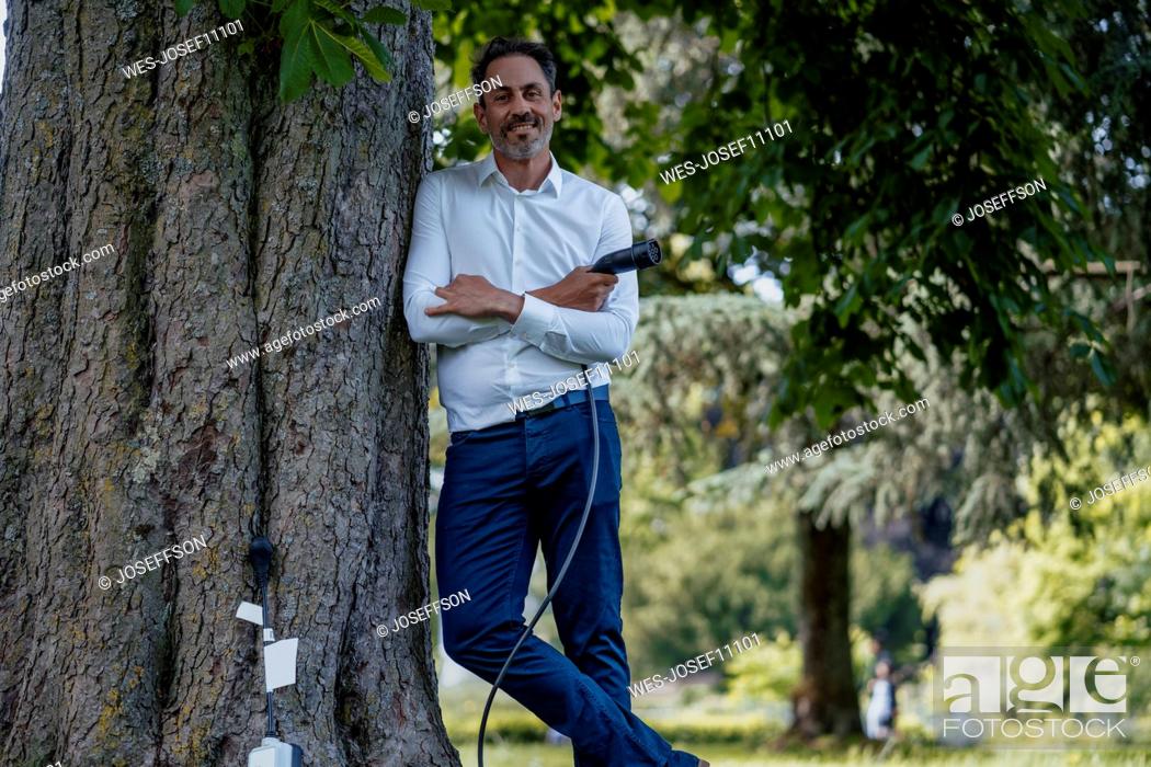 Photo de stock: Smiling mature businessman with arms crossed holding charging cable leaning on tree trunk.