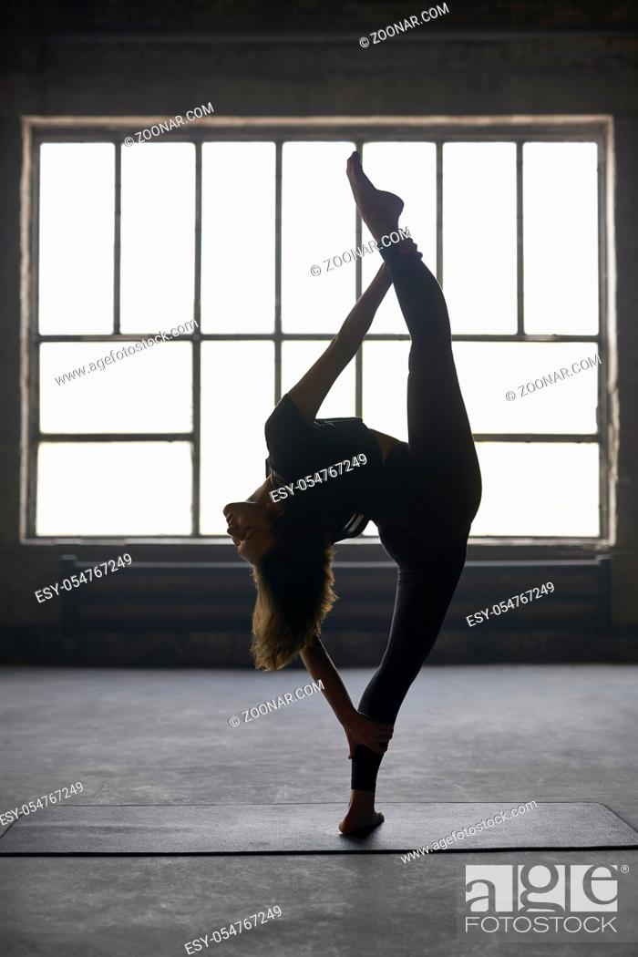 Stock Photo: Pretty girl in a black sportswear engaged in yoga on the window background in a loft style hall. She stands on the left leg on the mat and holds the right leg.
