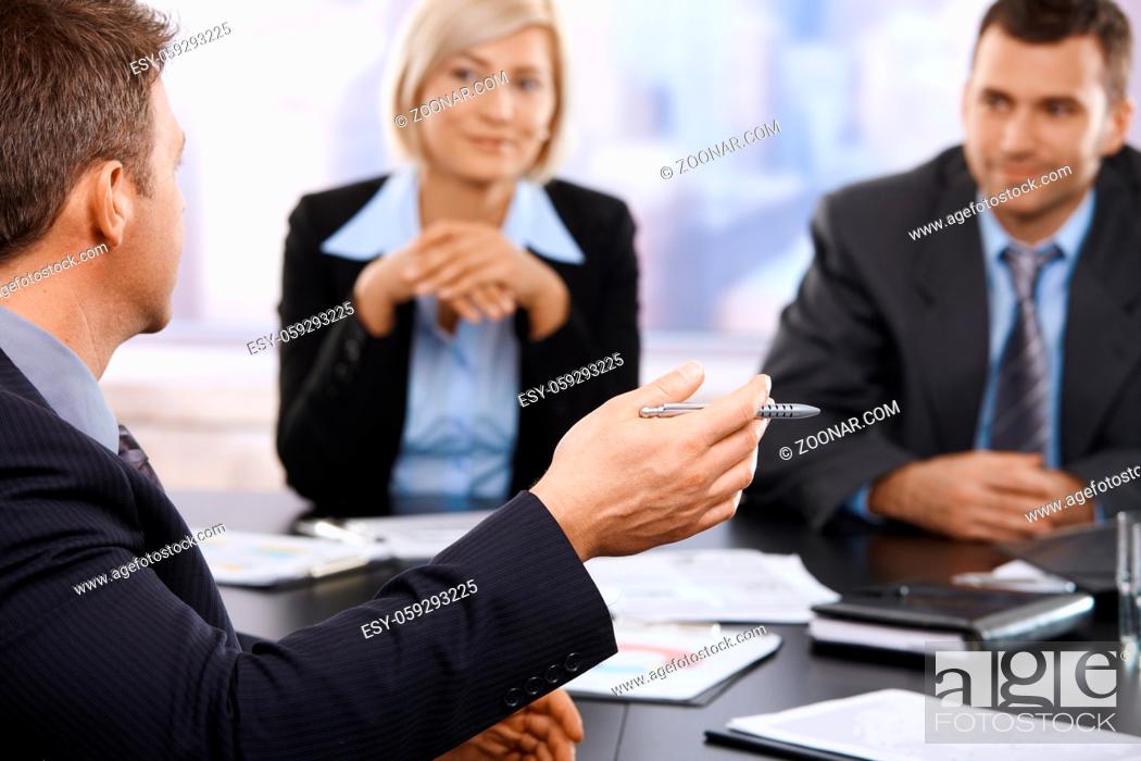 Stock Photo: Hand in closeup holding pen at business meeting, coworkers listening in the background. Focus on hand.