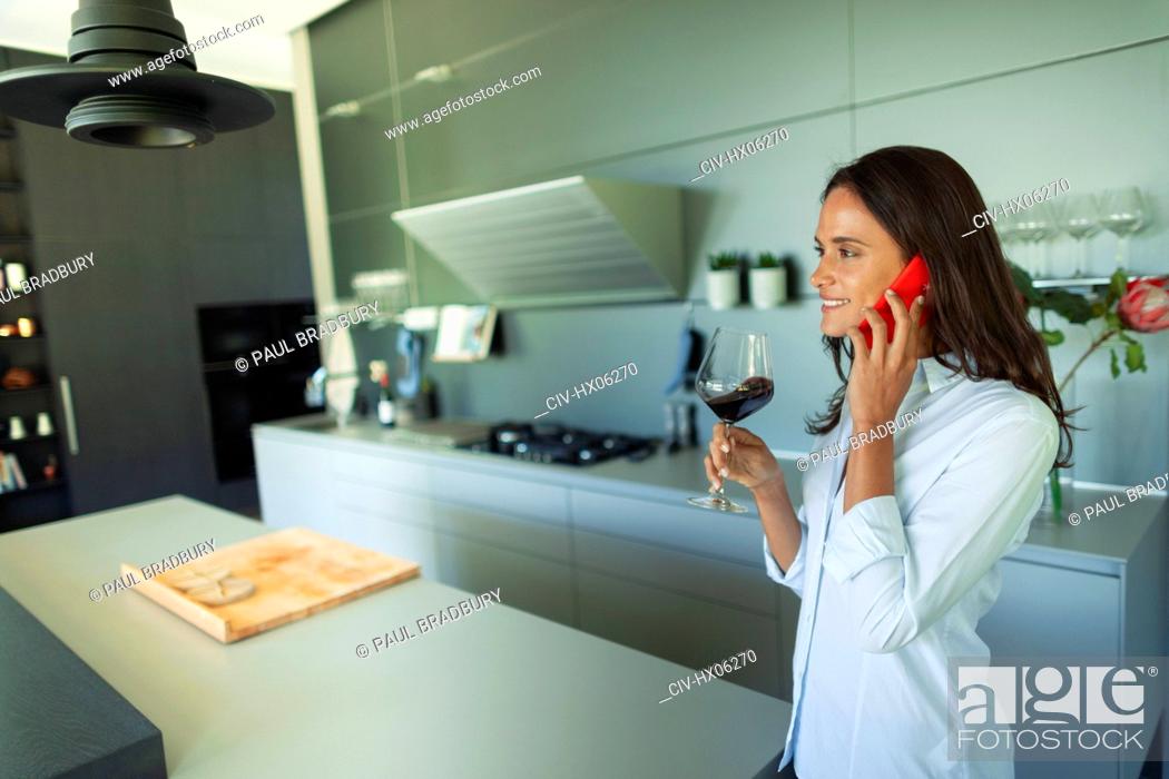 Stock Photo: Woman talking on smart phone and drinking red wine in modern kitchen.