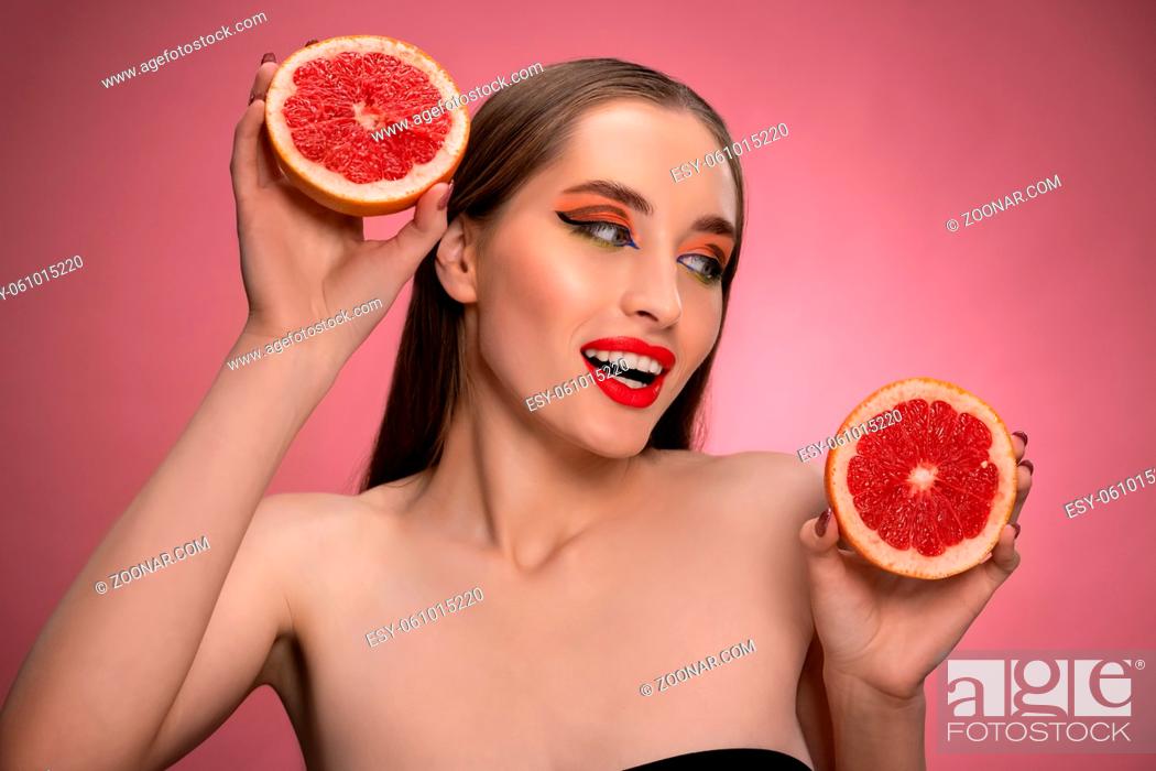 Stock Photo: Beautiful young woman model holding two juicy grapefruit sliced in half in her hands looking sideways. Charming joyful funny lady with red lips and long hair.