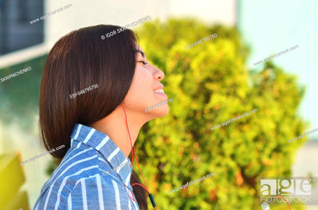 Stock Photo: Young beautiful is asian girl sitts on the bench near green tress and listen to music. Spring sunny day.