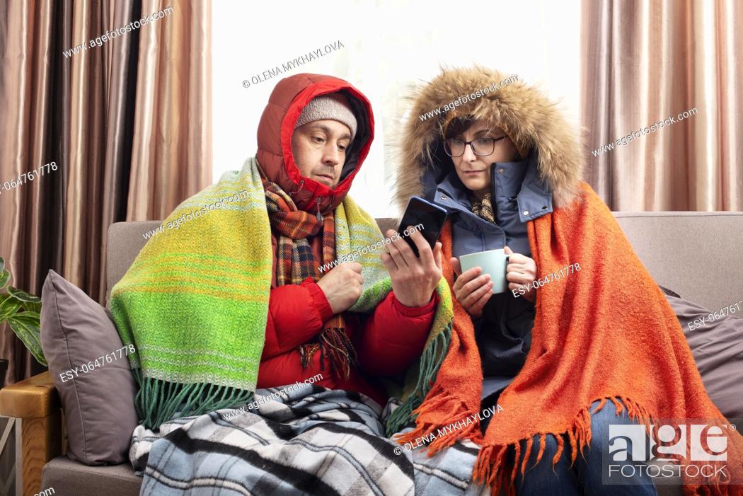 Imagen: Middle age family in parkas drinking coffee on the sofa while using smartphone in their cold living room.