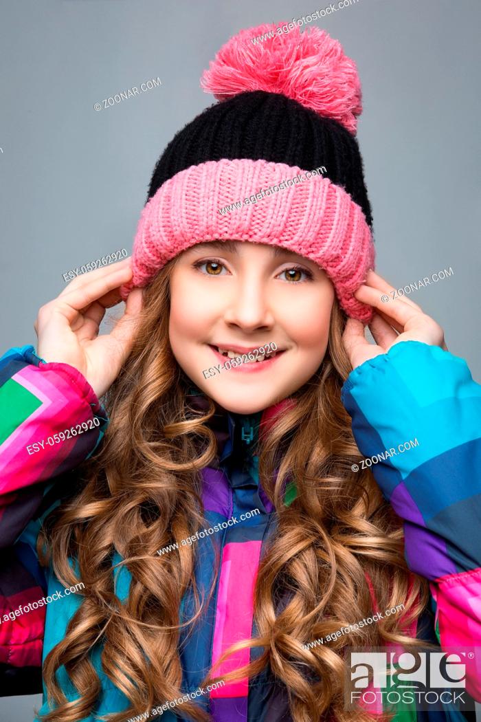 Stock Photo: Beautiful happy teen girl with long curly hair in pink wool hat and bright warm coat. Studio shot over grey background. Copy space.