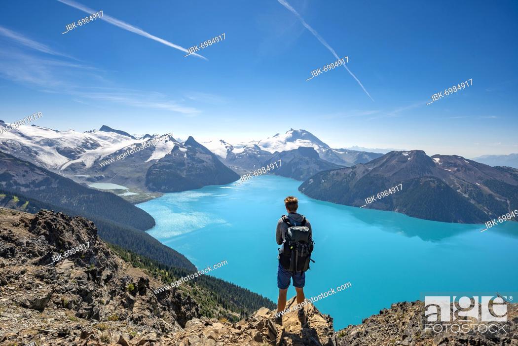 Photo de stock: Young man standing on a rock, looking into the distance, view of mountains and glacier with turquoise blue lake Garibaldi Lake, peaks Panorama Ridge.