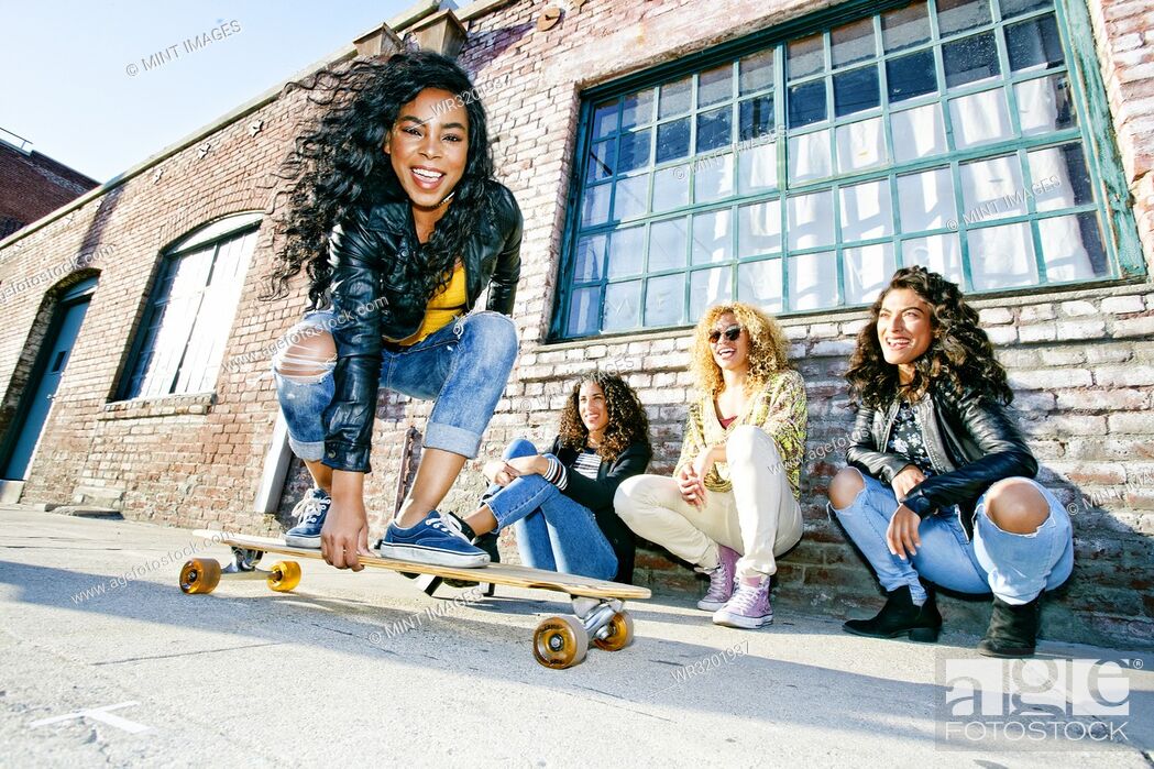 Stock Photo: Three young women with curly hair squatting in front of old building, watching smiling young woman riding a skateboard.
