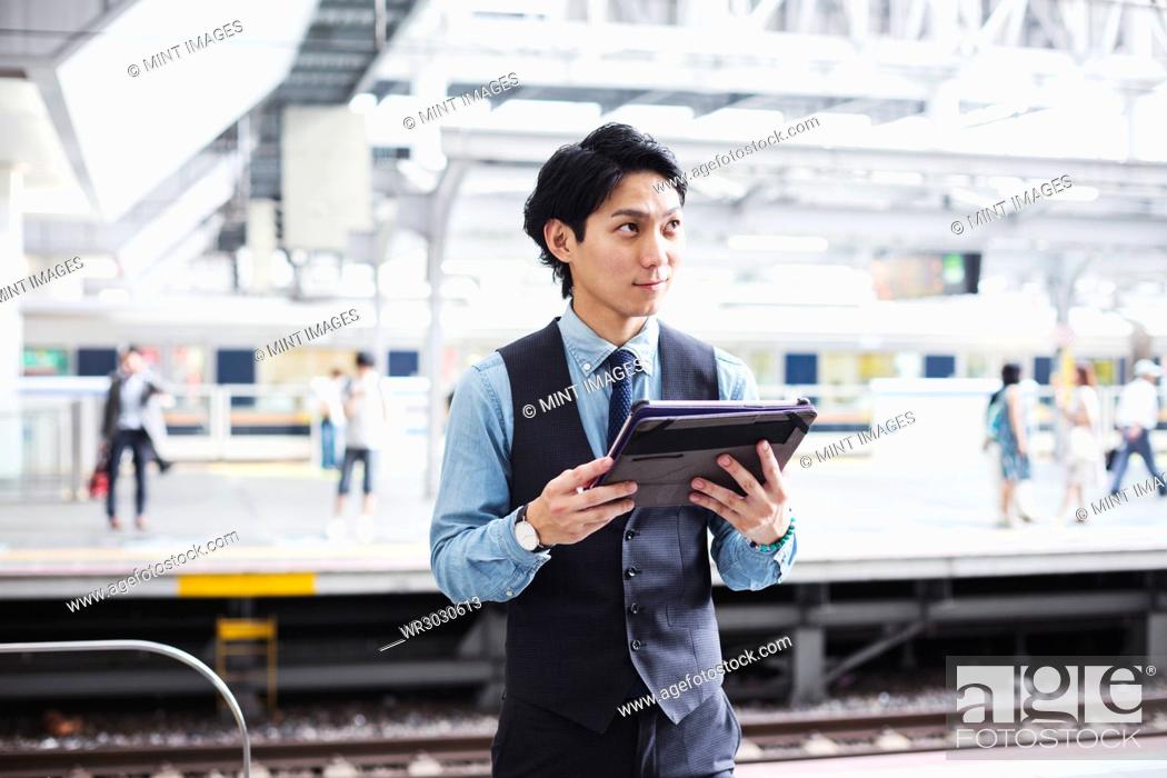 Stock Photo: Businessman wearing blue shirt and vest standing on train station platform, holding digital tablet.