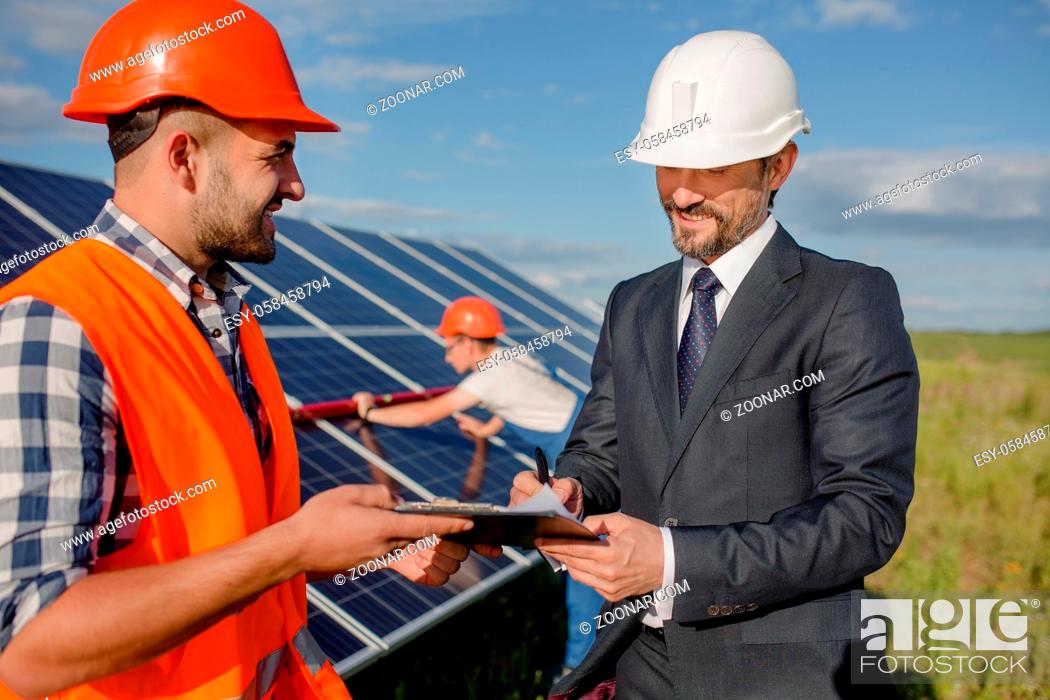 Stock Photo: Client signing agreement at solar energy station. Director and foreman signing papers while worker checking out solar panel behind them.