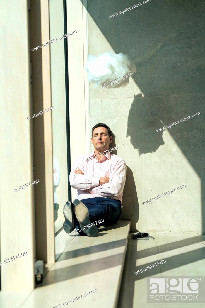 Stock Photo: Businessman with arms crossed sitting below cloud at window sill in office.