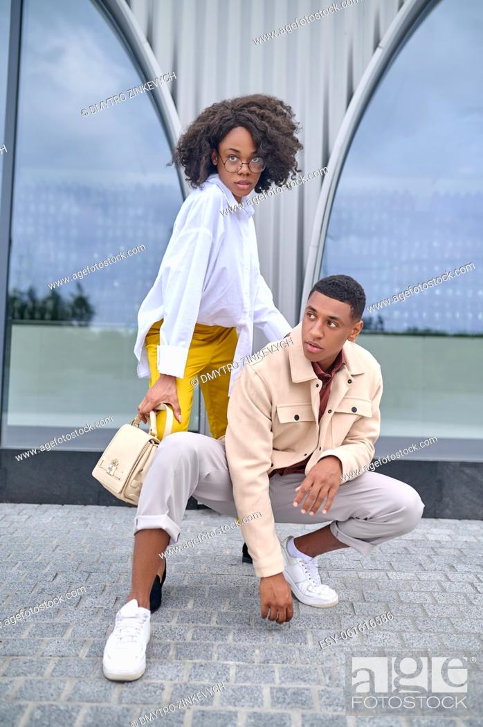 Stock Photo: Style. Stylish confident dark-skinned guy crouched and girl in glasses with bag posing on city street near building.