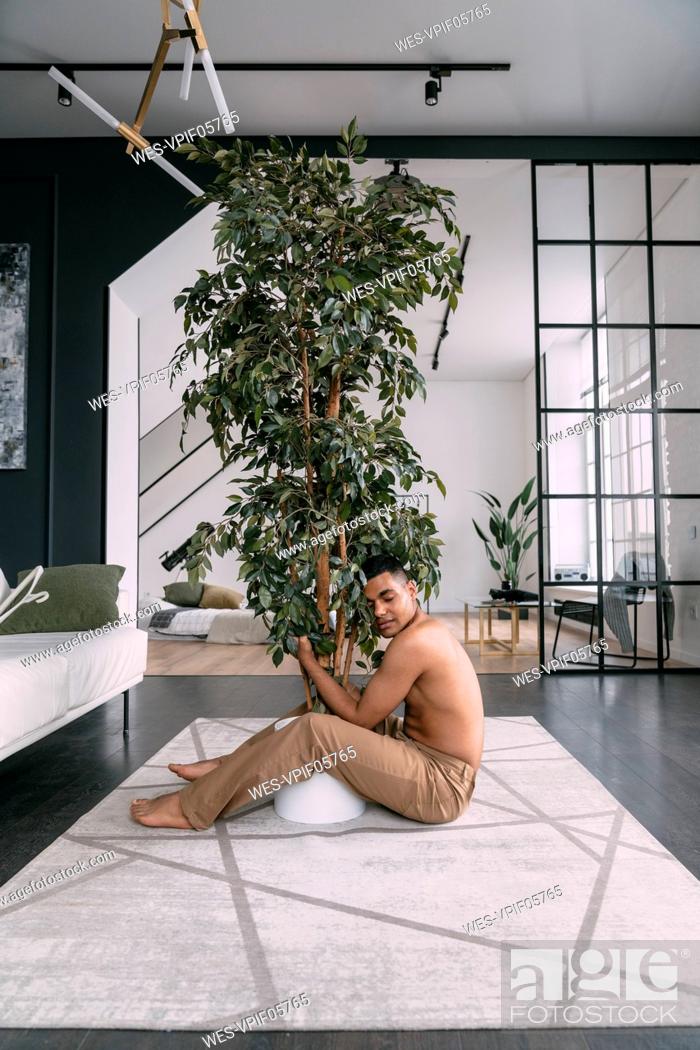 Stock Photo: Shirtless man with eyes closed embracing houseplant in living room at home.
