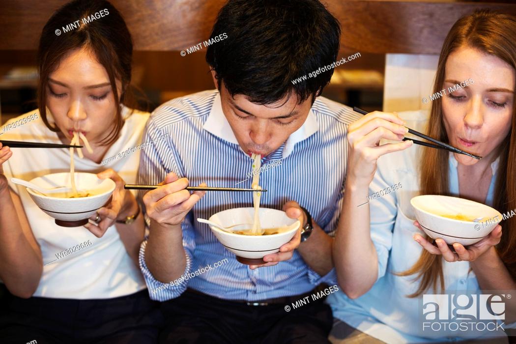 Stock Photo: High angle view of three people sitting sidy by side at a table in a restaurant, eating from bowls using chopsticks.