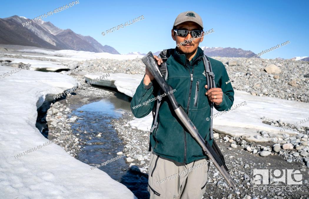 Stock Photo: 15 August 2019, Canada, Pond Inlet: The park ranger, Brian Koonoo, looks into the photographer's camera with his weapon on a hike to the glacier near Pond Inlet.