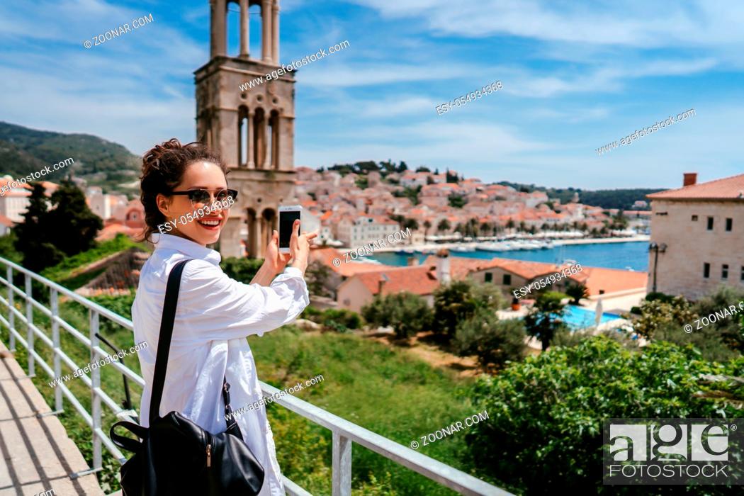 Stock Photo: Young beautiful woman posing for the camera on the background of a small Croatian town. Girl takes photo on smartphone.