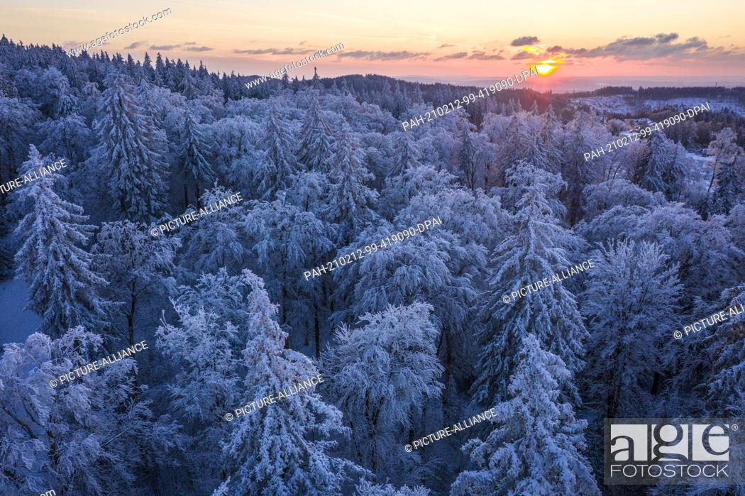 Stock Photo: 12 February 2021, Hessen, Sandplacken: The sun sets behind the trees covered with snow and ice at the Feldberg in Taunus (aerial view with a drone).