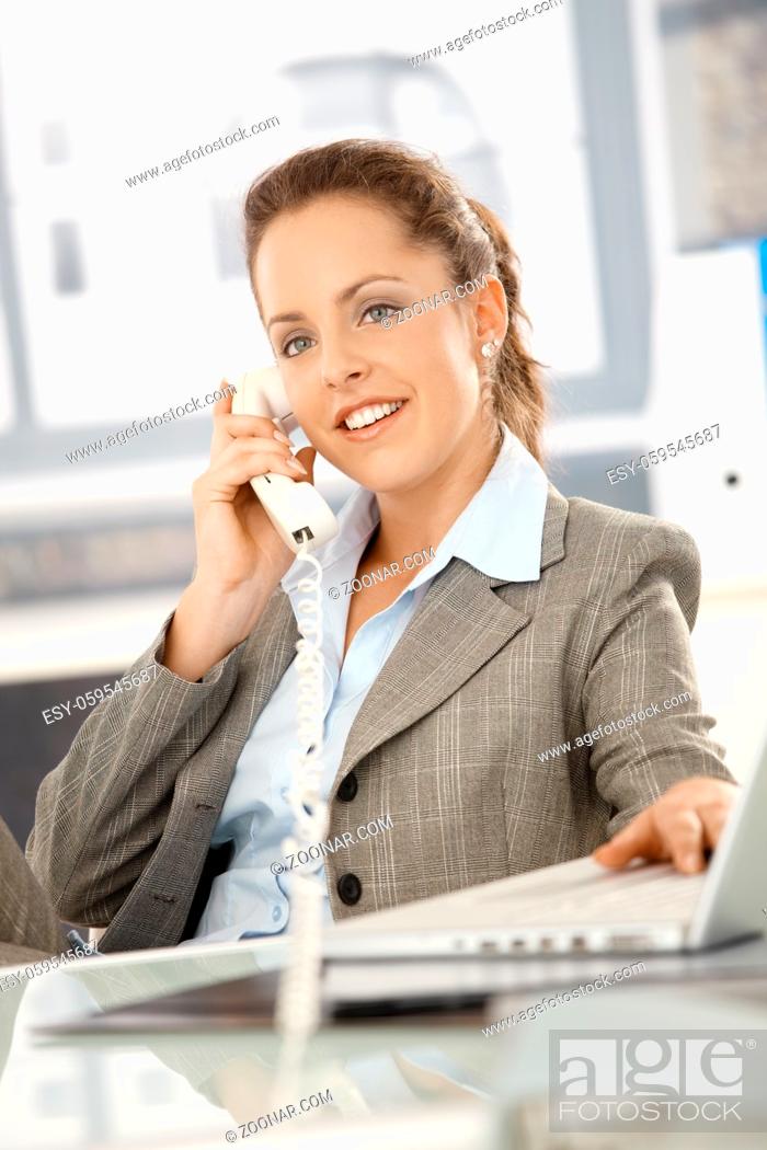 Stock Photo: Attractive businesswoman sitting at desk, talking on phone, having laptop, smiling.